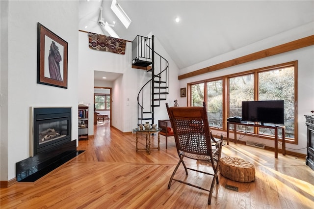 living area featuring a glass covered fireplace, ceiling fan, wood finished floors, stairs, and high vaulted ceiling