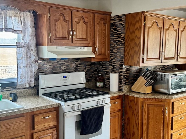 kitchen with tasteful backsplash, white range with gas stovetop, brown cabinets, and under cabinet range hood