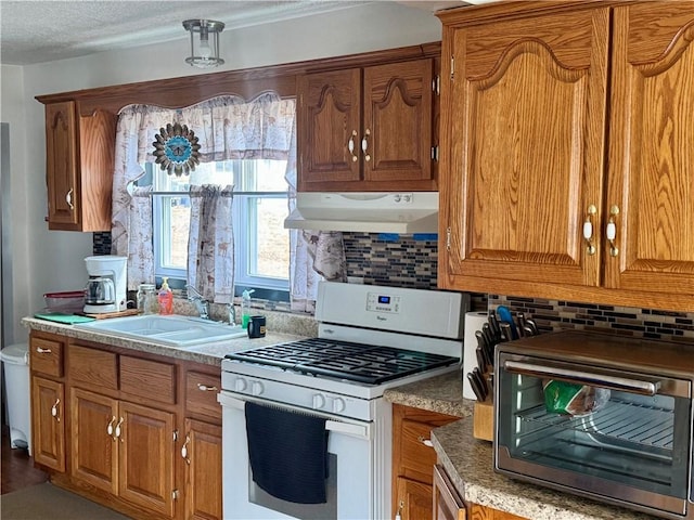 kitchen featuring white range with gas stovetop, under cabinet range hood, a sink, backsplash, and brown cabinetry