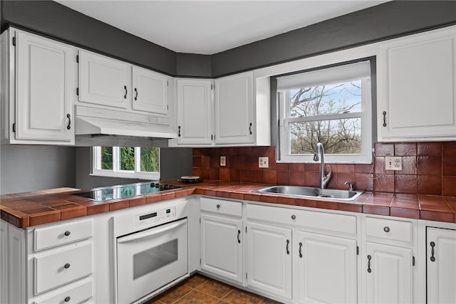 kitchen with tile counters, oven, black electric cooktop, under cabinet range hood, and a sink