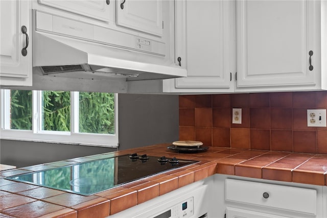 kitchen featuring tile countertops, black electric stovetop, under cabinet range hood, white cabinetry, and tasteful backsplash