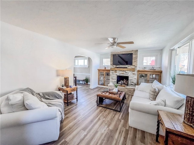 living room featuring arched walkways, a ceiling fan, a textured ceiling, a stone fireplace, and light wood-style floors