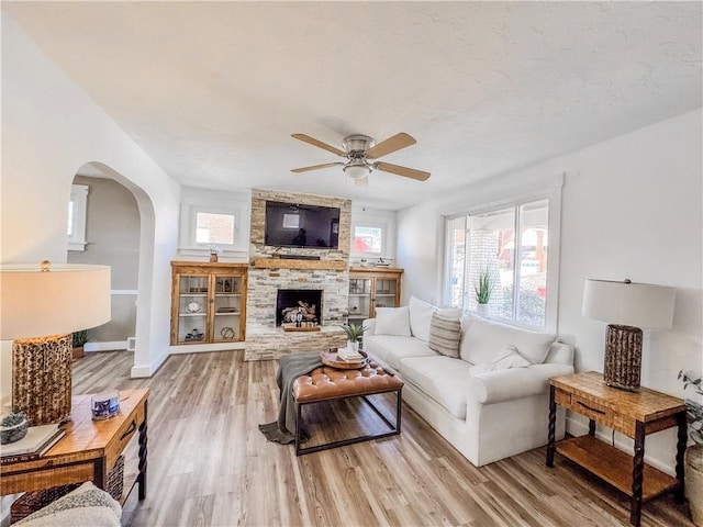 living room featuring arched walkways, ceiling fan, a fireplace, baseboards, and light wood-type flooring