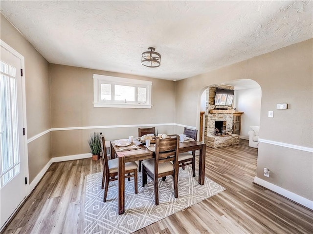 dining area featuring arched walkways, a stone fireplace, a textured ceiling, wood finished floors, and baseboards