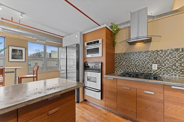 kitchen with brown cabinets, a warming drawer, decorative backsplash, appliances with stainless steel finishes, and island range hood