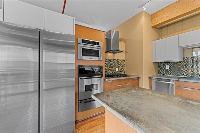 kitchen with light wood-style floors, appliances with stainless steel finishes, decorative backsplash, a warming drawer, and wall chimney exhaust hood