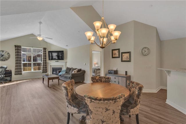 dining area with ceiling fan with notable chandelier, baseboards, vaulted ceiling, light wood-type flooring, and a glass covered fireplace