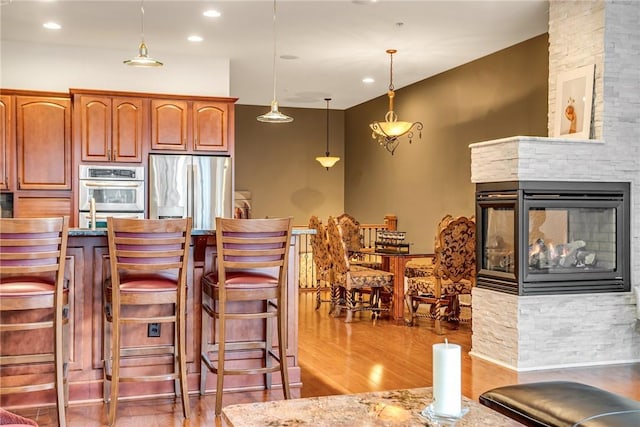 kitchen with a breakfast bar area, appliances with stainless steel finishes, wood finished floors, hanging light fixtures, and a stone fireplace