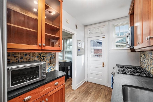 kitchen featuring brown cabinetry, decorative backsplash, light wood finished floors, stainless steel microwave, and glass insert cabinets