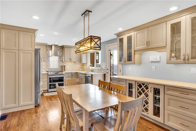 kitchen with stainless steel appliances, a sink, wall chimney range hood, light wood-type flooring, and decorative backsplash