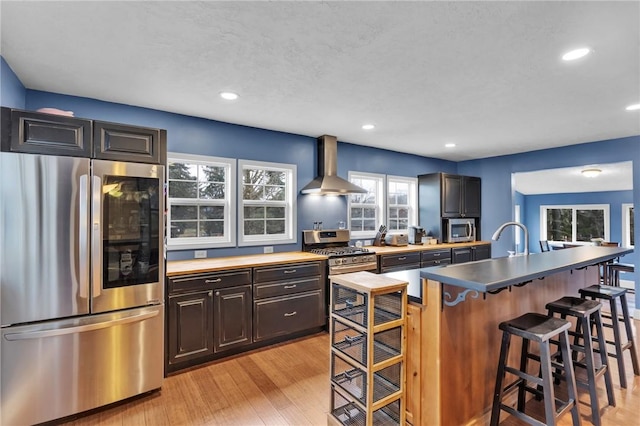 kitchen with a sink, a kitchen breakfast bar, appliances with stainless steel finishes, wall chimney range hood, and light wood finished floors