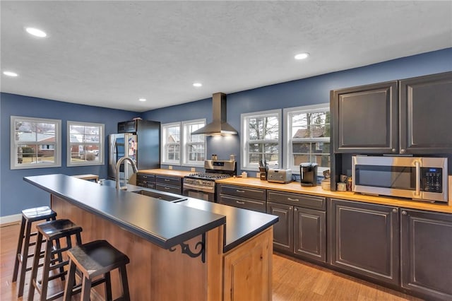 kitchen with a breakfast bar, light wood finished floors, appliances with stainless steel finishes, a sink, and wall chimney exhaust hood