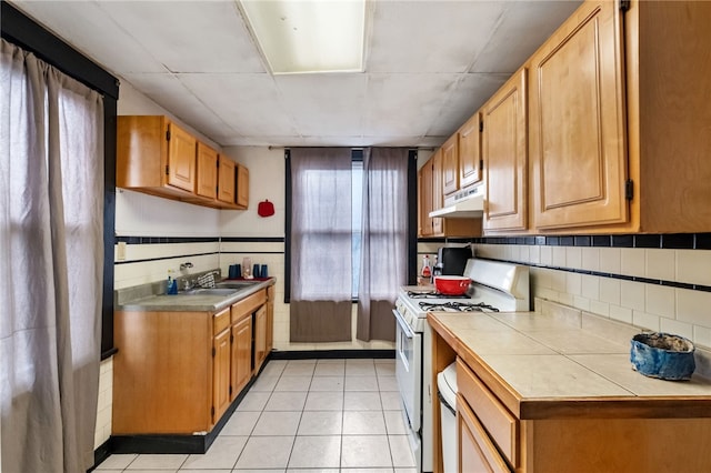 kitchen with light tile patterned floors, tile counters, white gas range, under cabinet range hood, and a sink