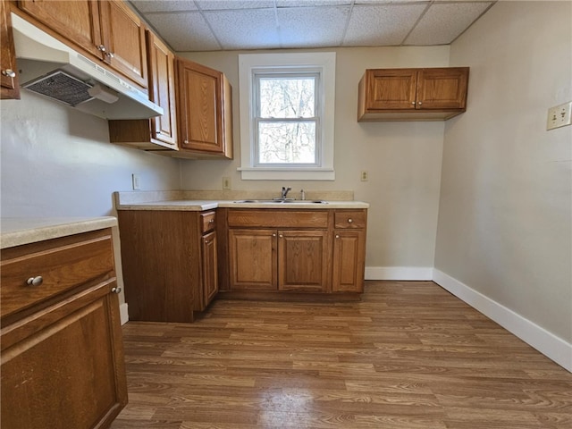 kitchen with baseboards, dark wood finished floors, brown cabinetry, under cabinet range hood, and a sink