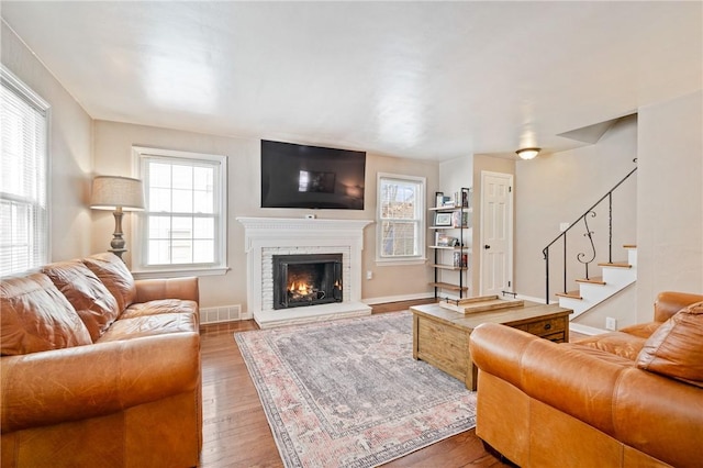 living room featuring wood finished floors, visible vents, baseboards, stairs, and a brick fireplace