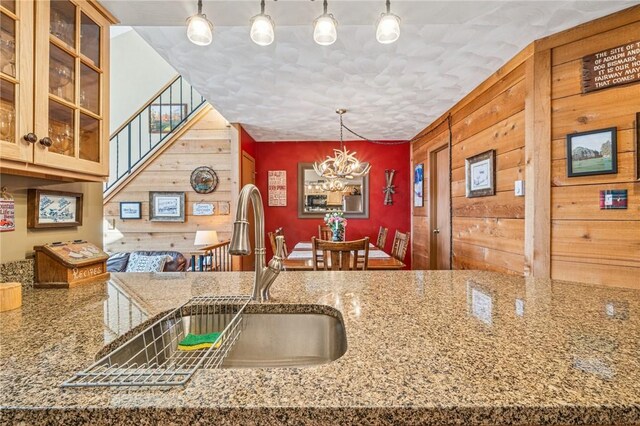 kitchen with glass insert cabinets, wood walls, a chandelier, pendant lighting, and a sink