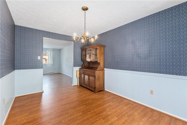 unfurnished dining area with wood finished floors, a wainscoted wall, an inviting chandelier, and wallpapered walls