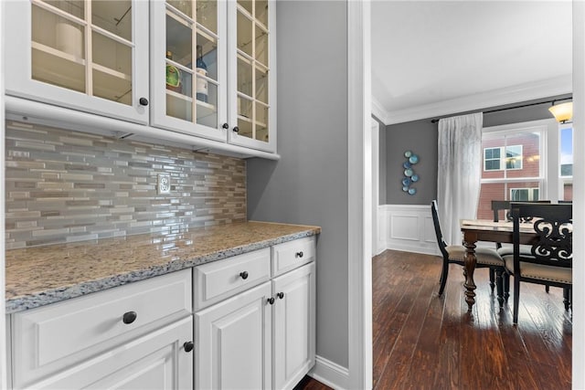 kitchen featuring dark wood-style flooring, crown molding, glass insert cabinets, wainscoting, and light stone countertops