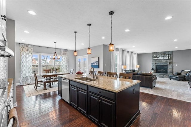 kitchen featuring dark wood finished floors, a kitchen island with sink, a sink, a stone fireplace, and dishwasher