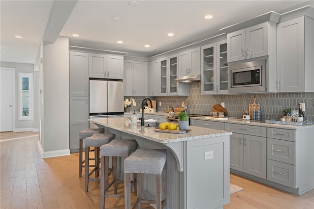 kitchen featuring under cabinet range hood, stainless steel appliances, gray cabinets, decorative backsplash, and light wood finished floors