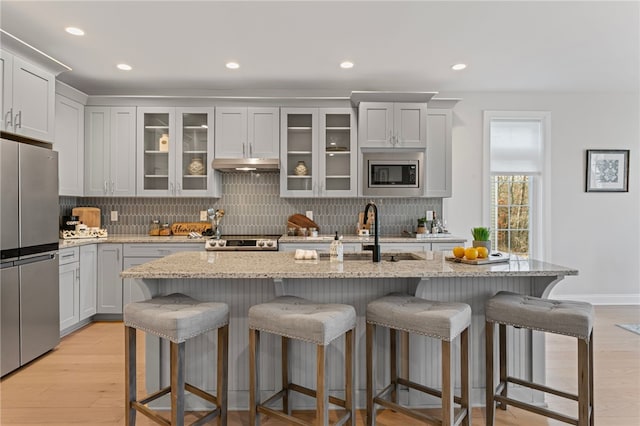 kitchen with light stone counters, under cabinet range hood, appliances with stainless steel finishes, decorative backsplash, and light wood finished floors