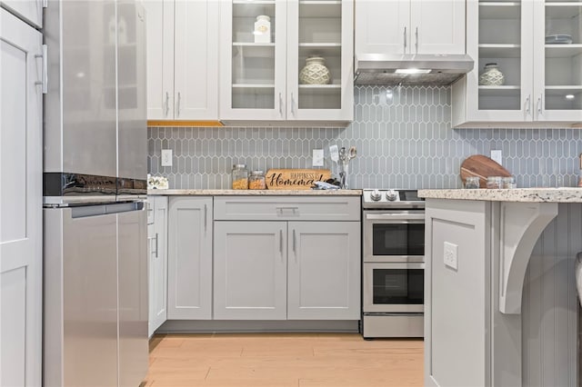 kitchen with under cabinet range hood, appliances with stainless steel finishes, backsplash, light stone countertops, and glass insert cabinets