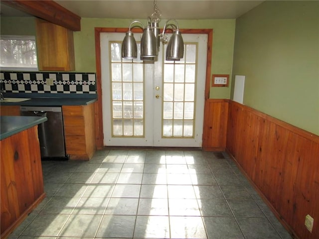 kitchen with a notable chandelier, stainless steel dishwasher, wainscoting, brown cabinetry, and dark countertops