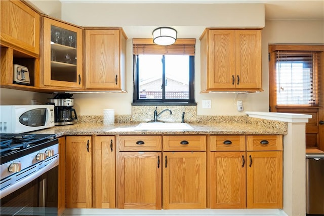 kitchen with stainless steel gas stove, white microwave, light stone counters, glass insert cabinets, and a sink