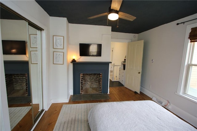 bedroom with wood-type flooring, a fireplace with raised hearth, ceiling fan, and baseboards