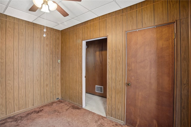 carpeted spare room with ceiling fan, wooden walls, visible vents, and a drop ceiling