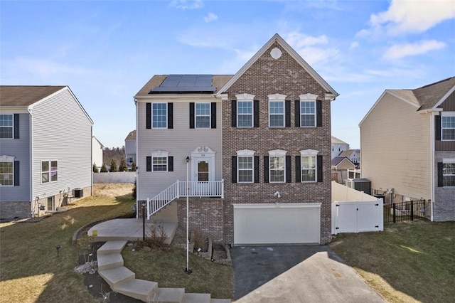 colonial-style house featuring roof mounted solar panels, brick siding, driveway, and fence