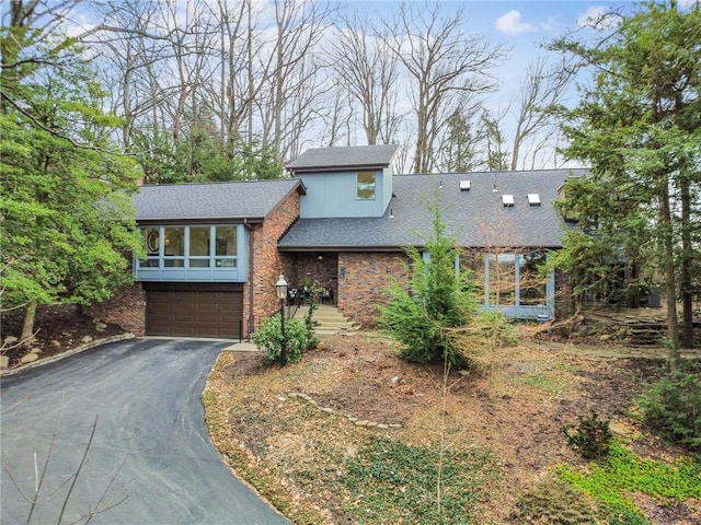 split level home featuring aphalt driveway, a garage, brick siding, a sunroom, and roof with shingles