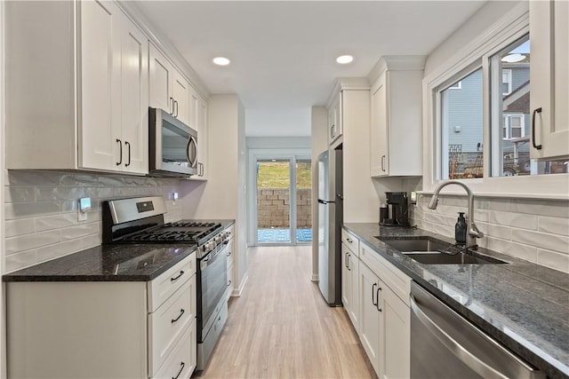 kitchen featuring light wood finished floors, white cabinets, appliances with stainless steel finishes, dark stone countertops, and a sink