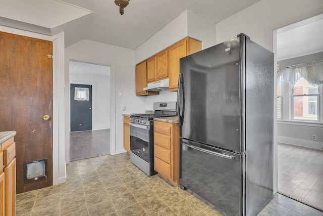 kitchen featuring under cabinet range hood, baseboards, freestanding refrigerator, brown cabinets, and stainless steel gas stove