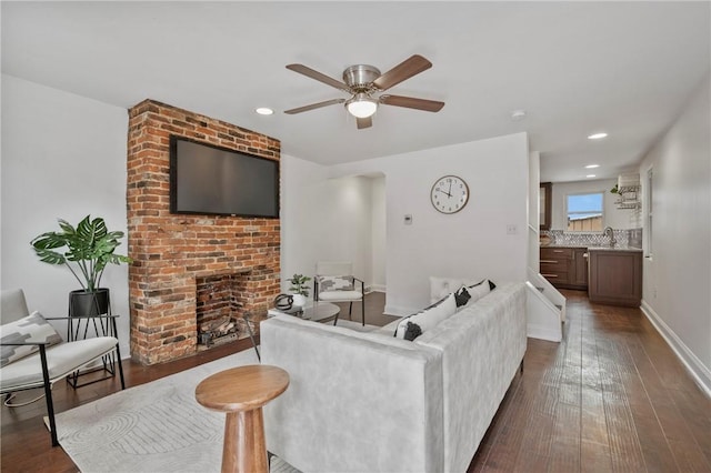 living room with baseboards, a ceiling fan, dark wood-style flooring, a brick fireplace, and recessed lighting