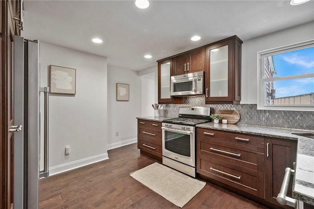 kitchen with light stone counters, dark wood-style flooring, appliances with stainless steel finishes, decorative backsplash, and glass insert cabinets