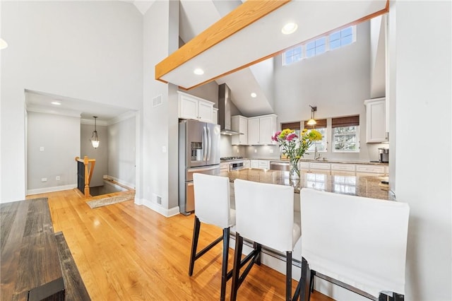 kitchen with light wood finished floors, stainless steel fridge, white cabinets, a breakfast bar area, and wall chimney range hood