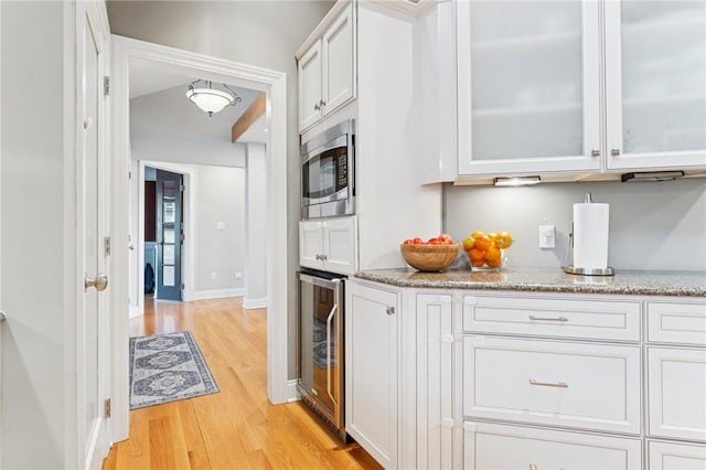 kitchen featuring wine cooler, light stone countertops, light wood finished floors, stainless steel microwave, and glass insert cabinets