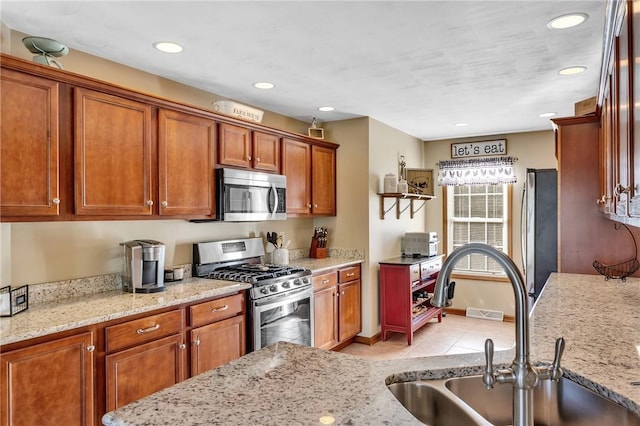 kitchen with appliances with stainless steel finishes, brown cabinets, light stone countertops, a sink, and light tile patterned flooring