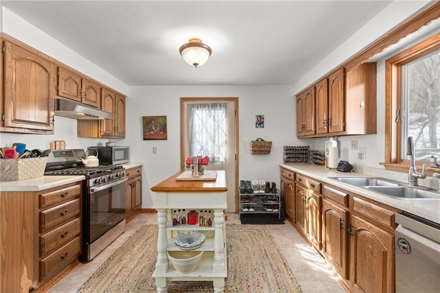 kitchen featuring under cabinet range hood, a sink, light countertops, appliances with stainless steel finishes, and brown cabinetry