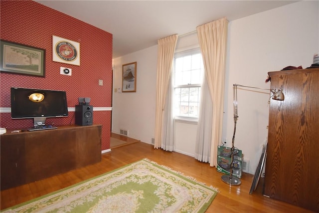 home office featuring wallpapered walls, an accent wall, visible vents, and wood finished floors