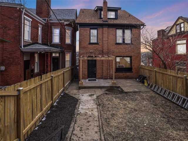 back of house at dusk featuring a patio, a fenced backyard, a chimney, roof with shingles, and brick siding