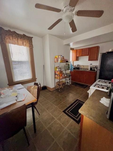 kitchen with white range with gas cooktop, dark tile patterned flooring, ceiling fan, brown cabinets, and freestanding refrigerator