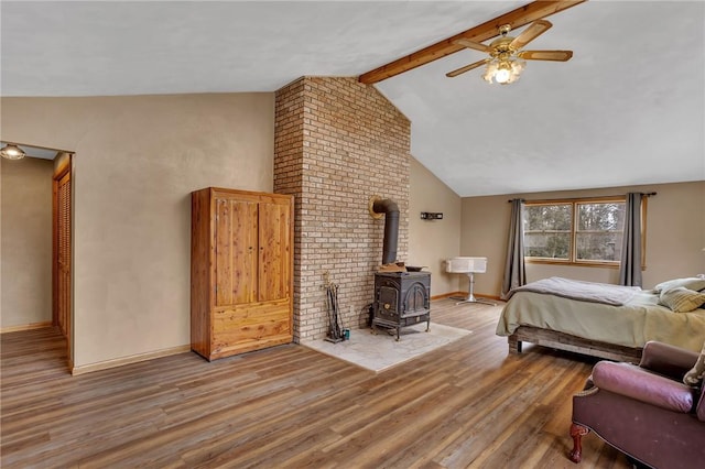 bedroom with baseboards, ceiling fan, wood finished floors, a wood stove, and vaulted ceiling with beams