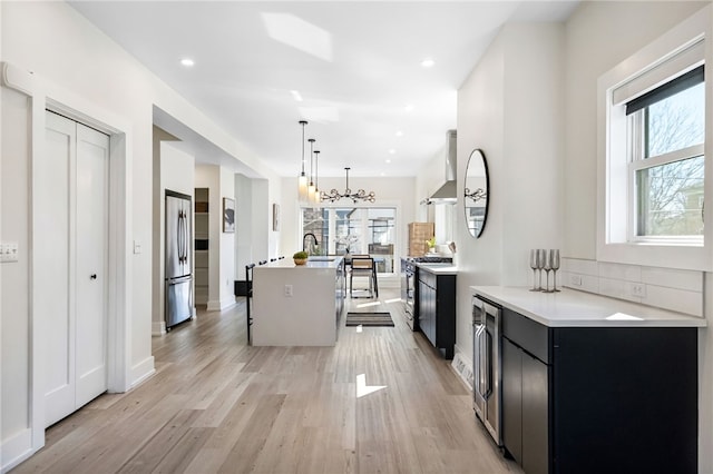 kitchen with appliances with stainless steel finishes, wall chimney range hood, an island with sink, light wood-type flooring, and dark cabinetry