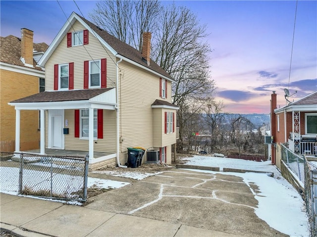 snow covered property with roof with shingles, a chimney, central air condition unit, a porch, and fence