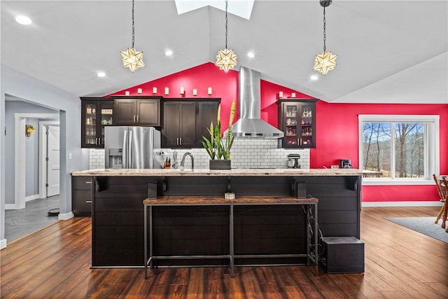 kitchen featuring dark wood-style flooring, a center island with sink, stainless steel fridge with ice dispenser, wall chimney range hood, and a kitchen bar