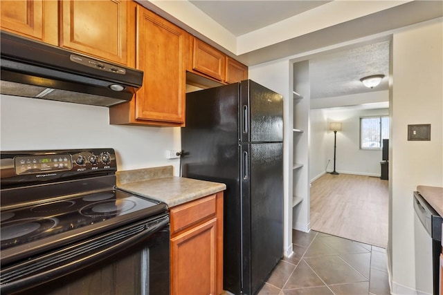 kitchen featuring dark tile patterned flooring, brown cabinets, under cabinet range hood, light countertops, and black appliances