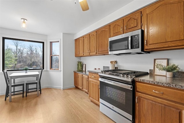 kitchen with light wood finished floors, stainless steel appliances, brown cabinetry, dark stone countertops, and baseboards