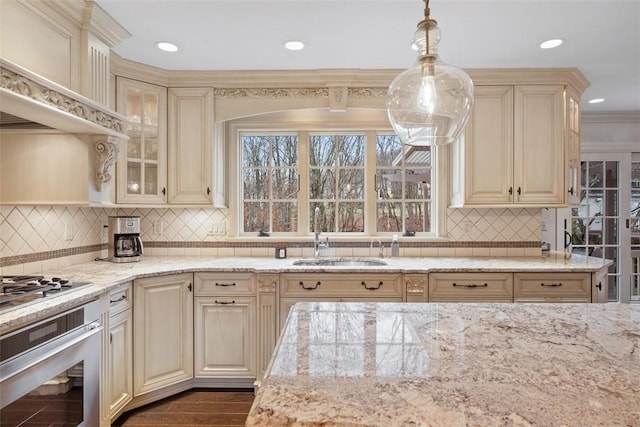 kitchen featuring cream cabinets, stainless steel appliances, a sink, hanging light fixtures, and light stone countertops